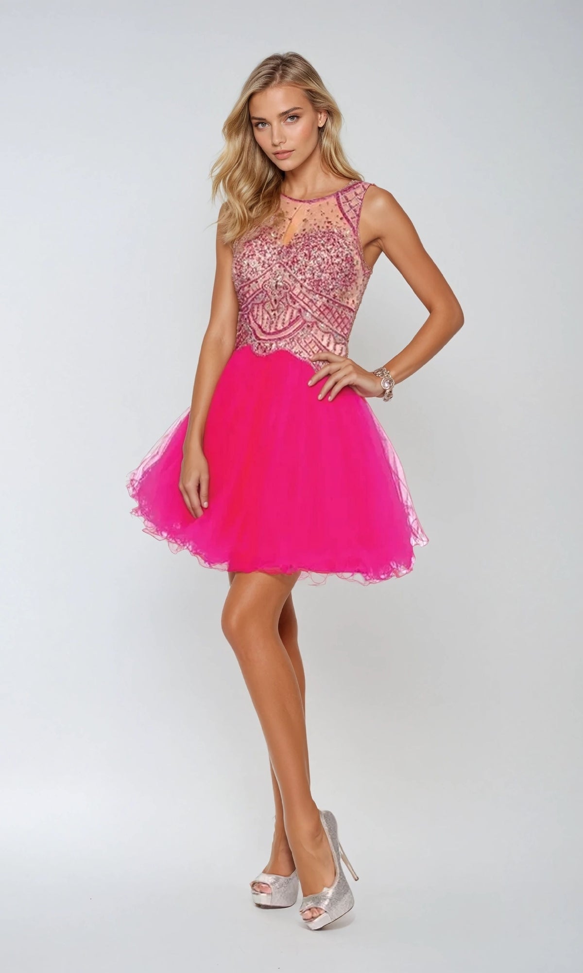 A woman poses in the Juliet 761 short party dress—a hot pink, sleeveless mini dress with a beaded bodice, sheer neckline, and A-line tulle skirt—paired with silver high heels against a light background. Perfect for homecoming!.